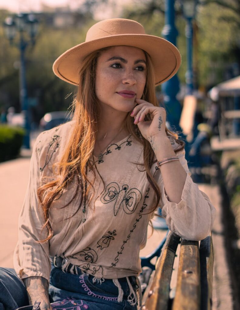 Woman in a chic hat and embroidered blouse sits on a park bench enjoying a sunny day.