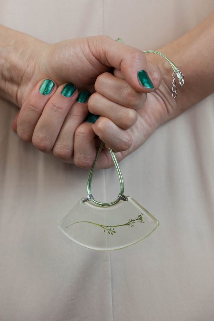 A close-up shot of hands with green nails holding a delicate pendant necklace indoors.