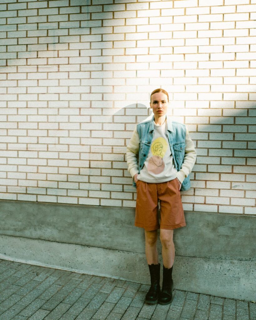 Model in blue vest and shorts posing against brick wall in sunlight.