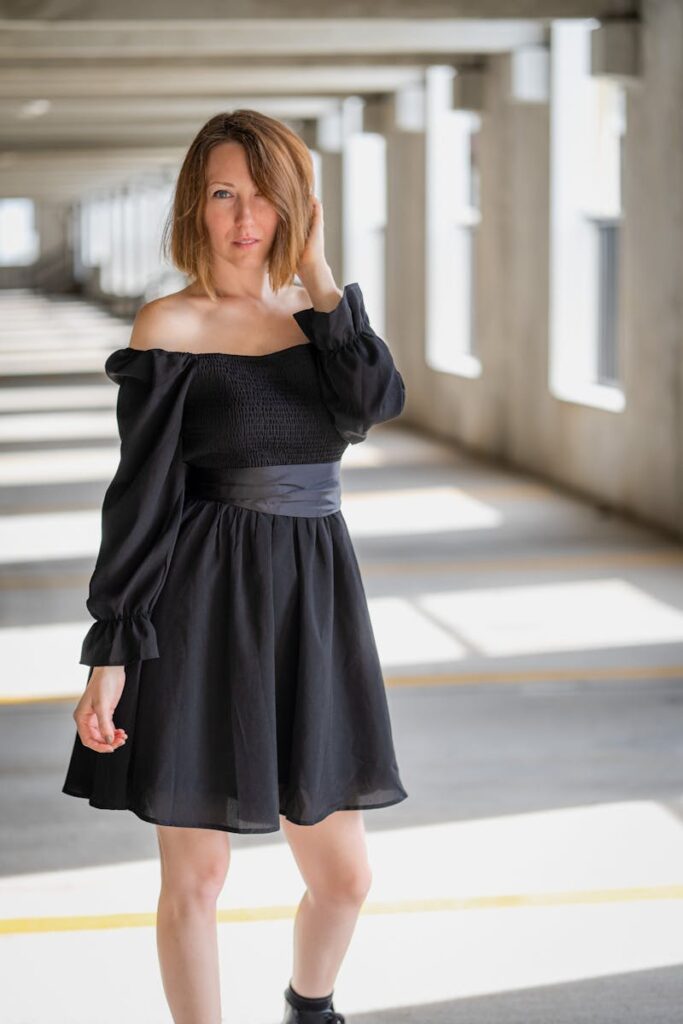 Elegant woman in black dress poses confidently in a sunlit corridor.