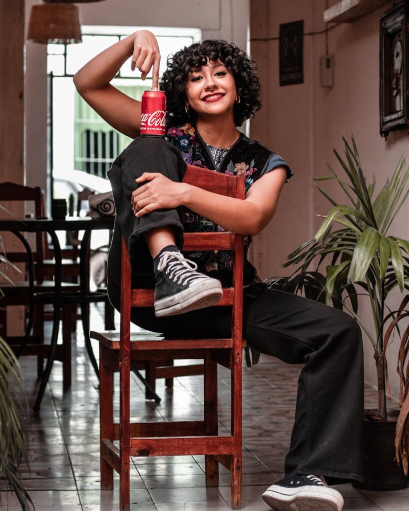 Hispanic woman with curly hair sits smiling, posing with a soda can inside a cafe.