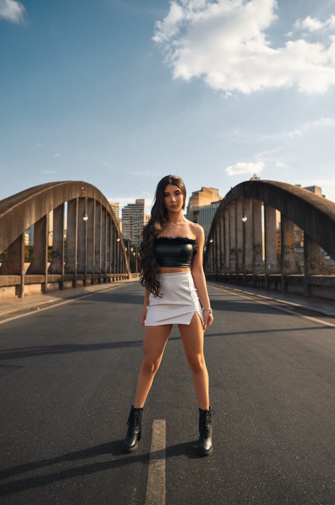 Fashionable woman standing confidently on a bridge with city skyline in the background during sunset.