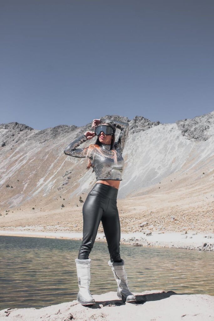 A woman in a silver sci-fi outfit poses by a serene mountain lakeshore under a clear sky.