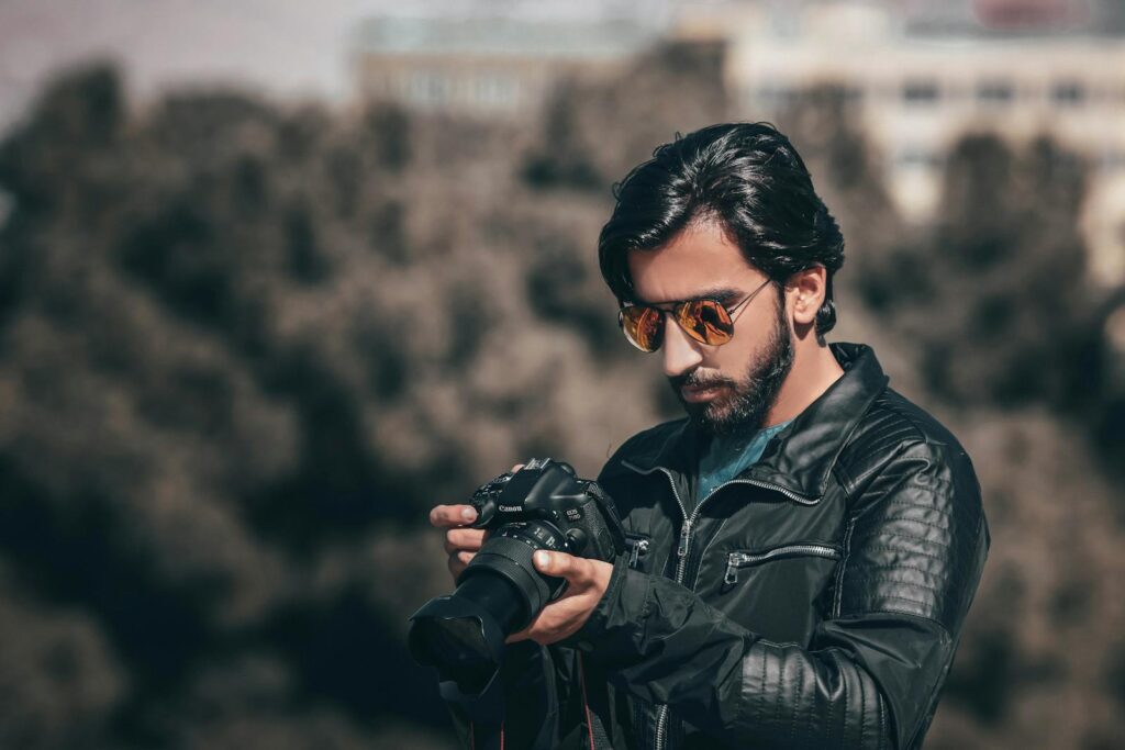 Portrait of a stylish male photographer in sunglasses examining a DSLR camera outdoors.