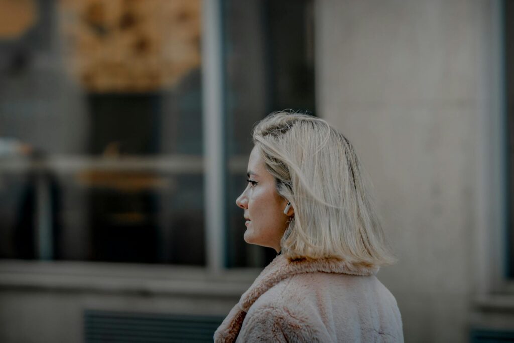 Profile of a stylish blonde woman walking outdoors in an urban environment, wearing a coat.