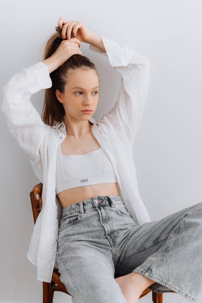 Woman in casual fashion sitting and adjusting her hair against a white background.