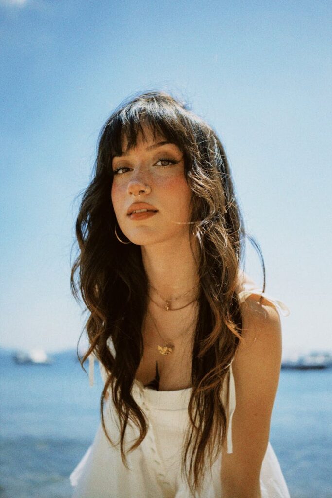 A woman with flowing hair and a white dress gazes calmly by the sea on a sunny day.