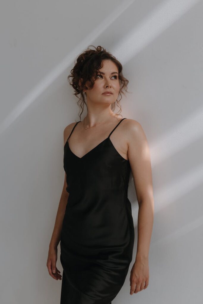 Stylish woman in a black dress poses gracefully against a softly lit wall, embodying elegance and simplicity.