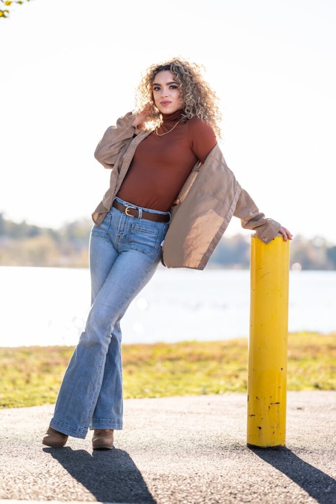 Fashion-forward woman with curly hair posing by a lake on a sunny day in casual attire.