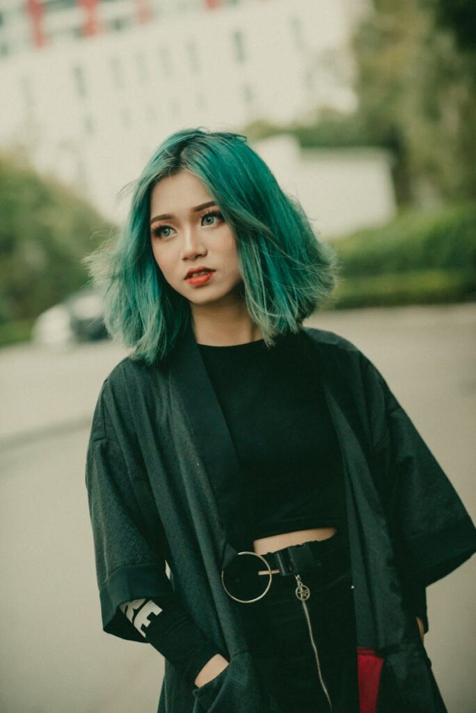 A fashionable young woman with teal hair poses outdoors in an urban area, displaying a modern and stylish look.