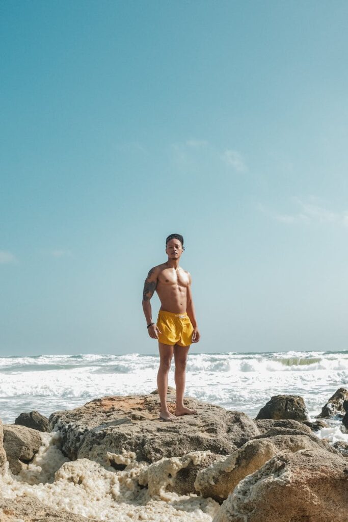 Man in yellow shorts standing on rocks with ocean waves in the background.