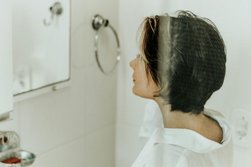 Profile view of woman with short hair looking in a bathroom mirror, illustrating a personal hygiene and reflection moment.