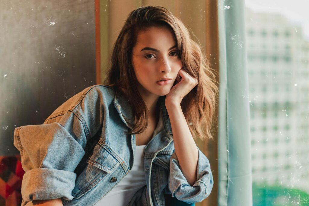 Young woman posing thoughtfully by a window wearing a denim jacket.