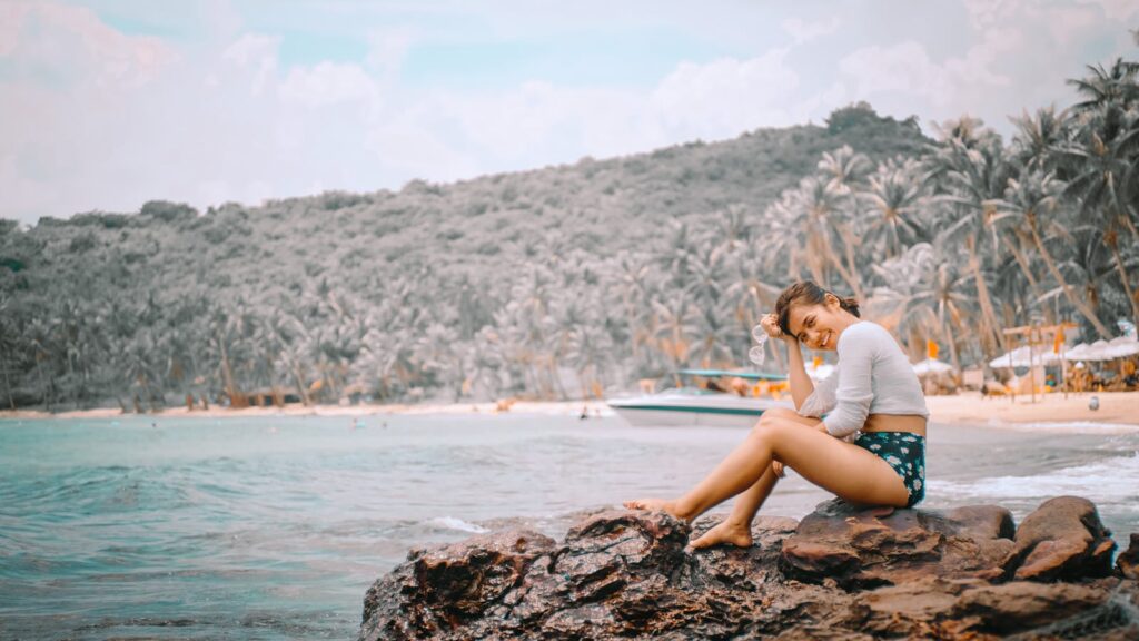 A woman enjoying a sunny day on a tropical beach, sitting on a rock by the sea.