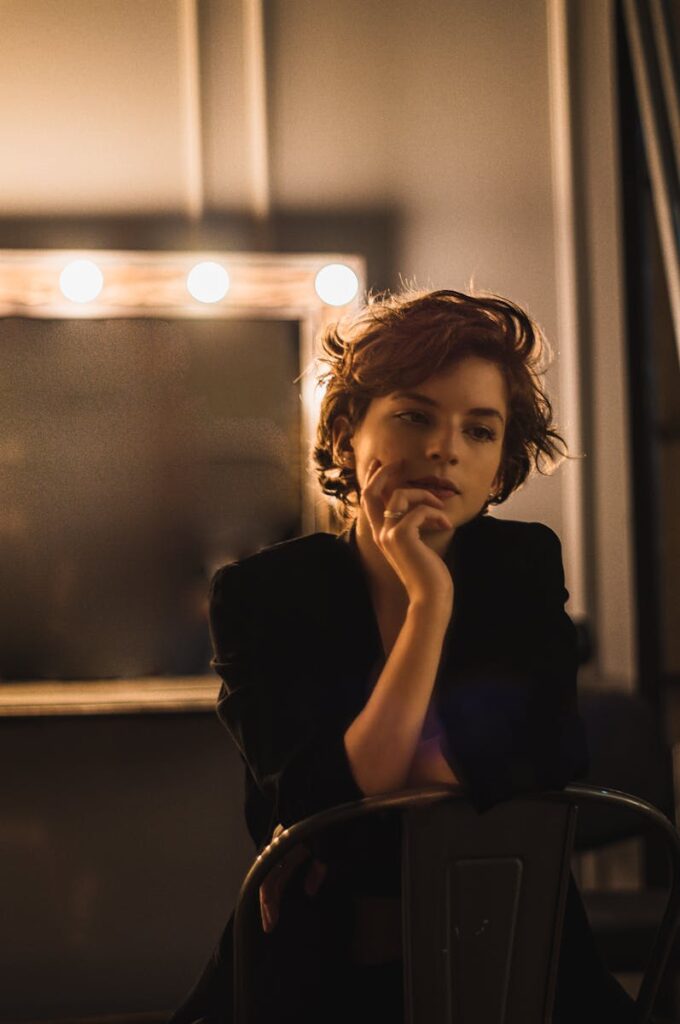 A woman with short hair sits pensively in a dimly lit dressing room, reflecting thoughtful elegance.