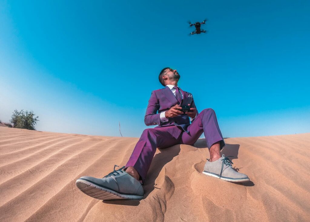 A stylish man in a suit controls a drone against a clear blue sky in the Dubai desert.