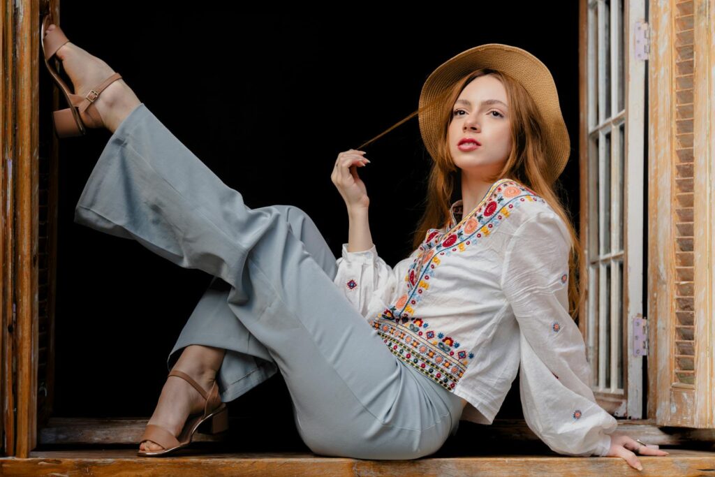 Young woman posing elegantly by a windowsill, wearing fashionable embroidered blouse and hat.