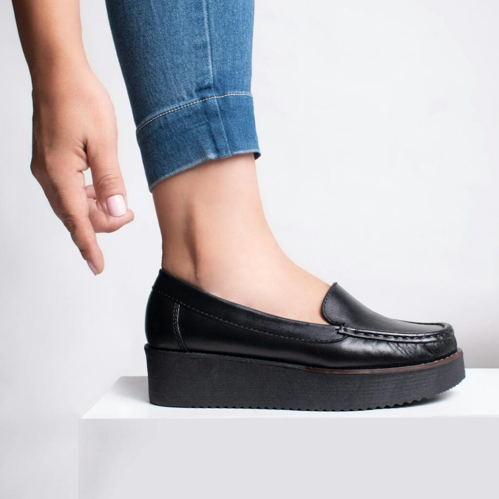 Close-up of a leg wearing a black platform loafer with a hand adjusting the shoe, captured in a studio setting.