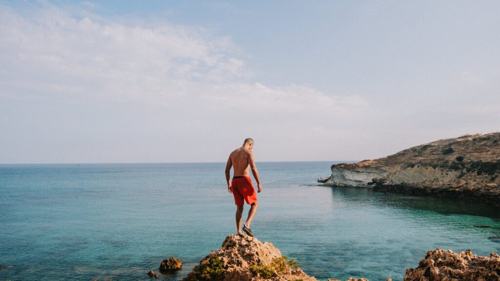 A man in swim trunks admires the seascape while standing on rocky cliffs.