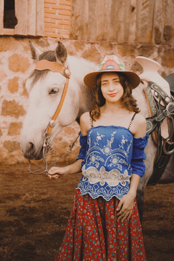 A young woman stands with a horse, wearing colorful traditional attire in a rustic setting.