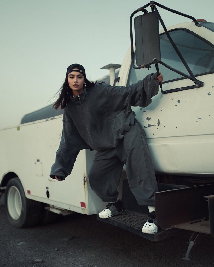 Stylish woman in urban streetwear poses by a truck at dusk, exuding modern fashion vibes.