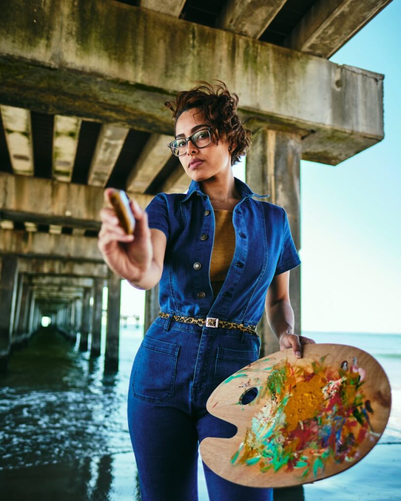 Stylish female artist in blue jumpsuit painting under urban pier with ocean backdrop.