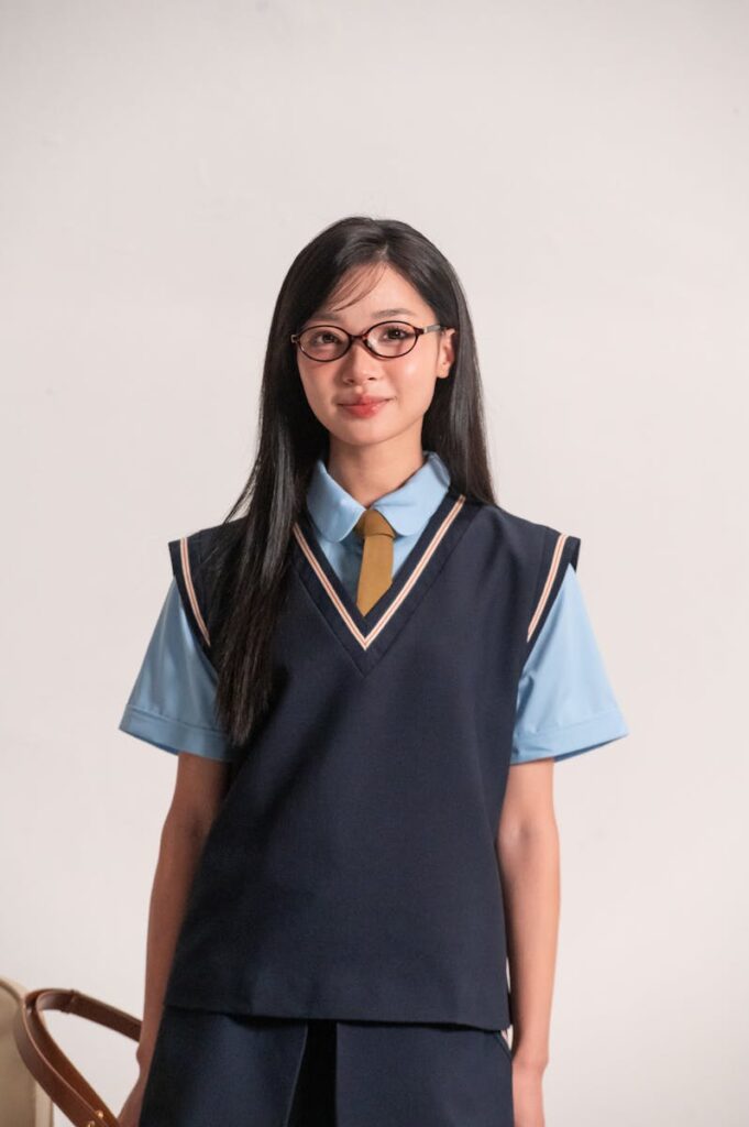 Portrait of a young adult woman wearing a school uniform and glasses, standing indoors.