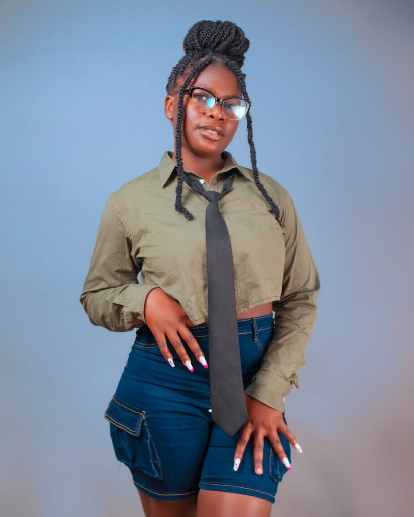 A young woman poses confidently in casual attire with glasses, braids, and a tie.