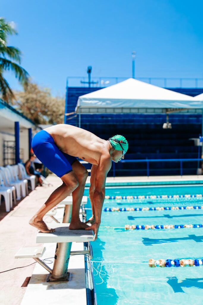 Male swimmer in blue shorts poised to dive at an outdoor pool under a clear sky.