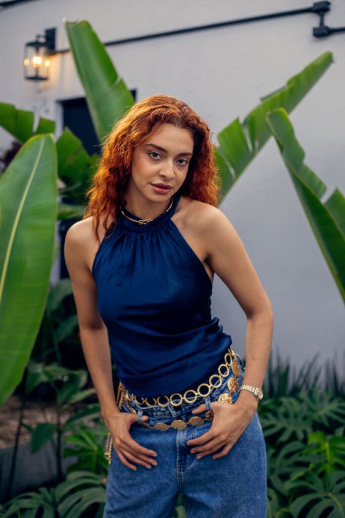 Woman with red hair in a blue top stands amidst lush tropical leaves.