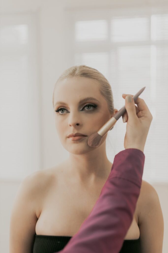 Close-up of a makeup artist applying blush to a young woman indoors.
