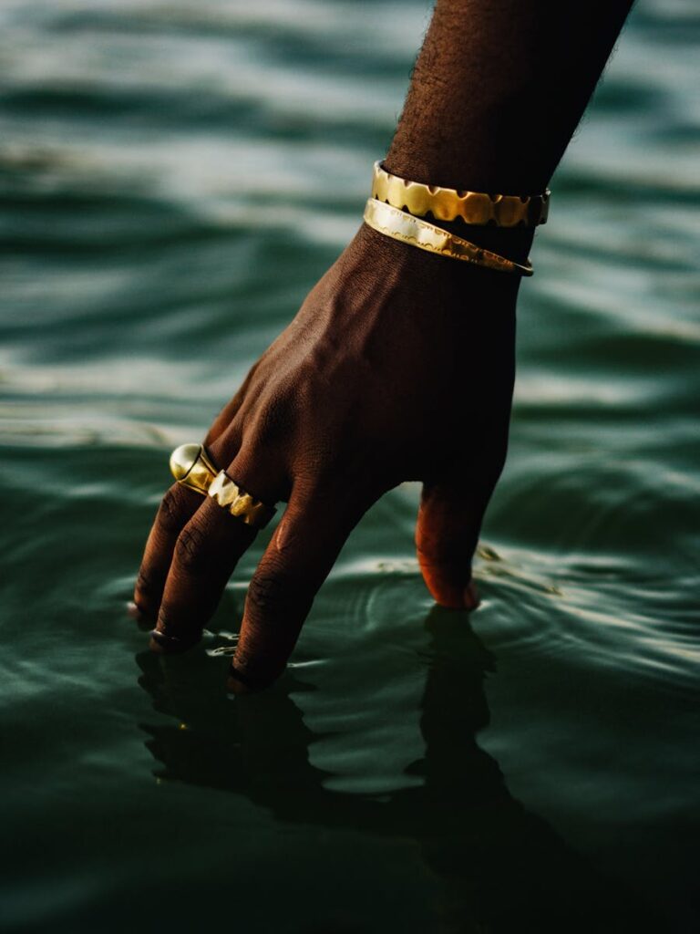 A hand adorned with gold jewelry gently touches calm water, creating a serene and luxurious atmosphere.