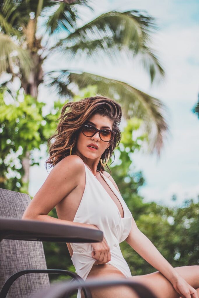 Stylish woman in a white swimsuit posing with sunglasses at a tropical resort.