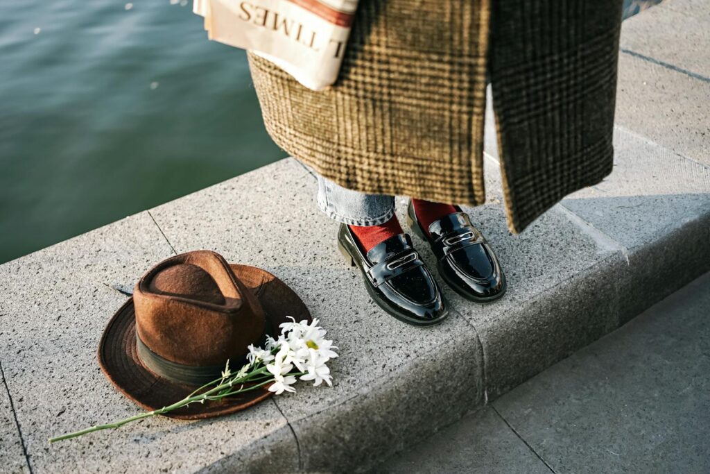 Chic fashion ensemble featuring patent loafers, hat, and newspaper by the water.