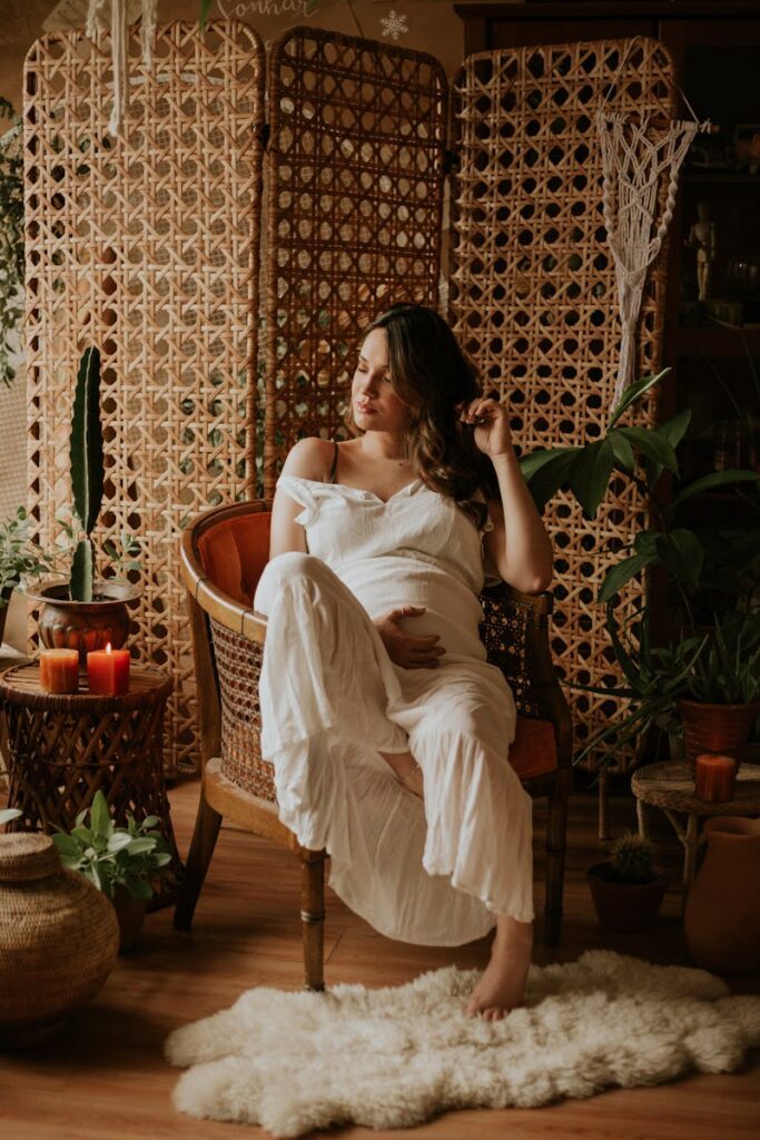 Pregnant woman in a white dress sitting in a cozy indoor space with plants and warm lighting.