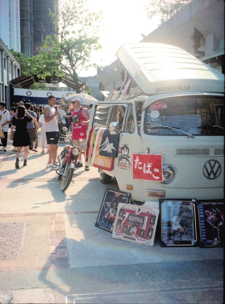 Lively vintage flea market scene in Taipei with a Volkswagen van and diverse crowd on a sunny day.