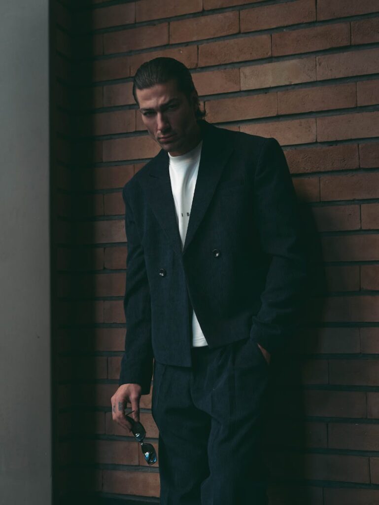 Man in elegant dark suit stands against brick wall holding sunglasses.