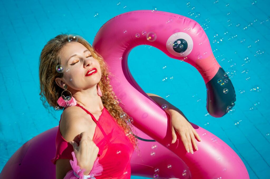 Young woman enjoying sun in a pool with vibrant pink flamingo float and bubbles.