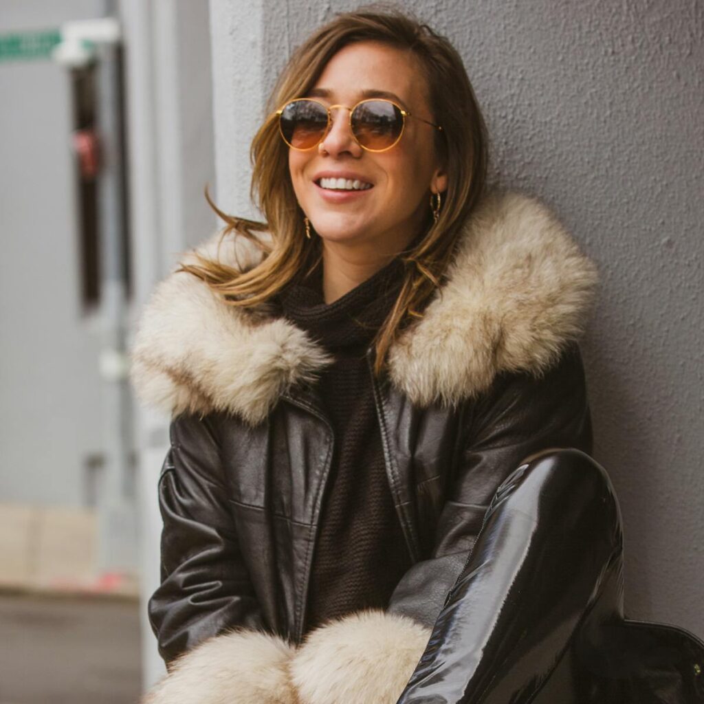 Fashionable woman with sunglasses and fur-lined jacket smiling outdoors by a wall.