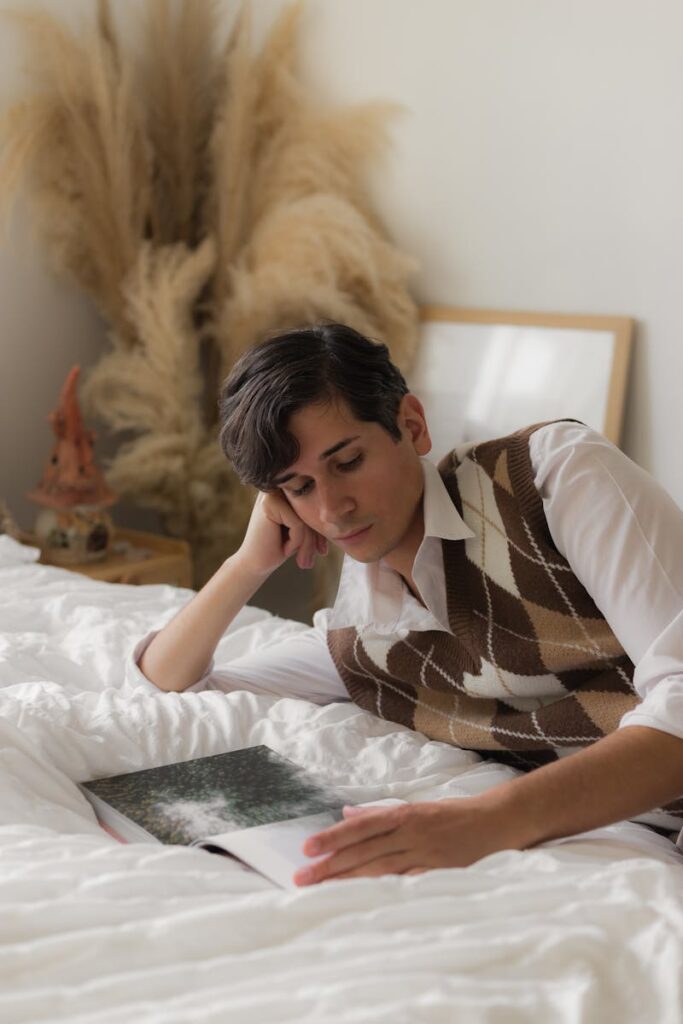 A young man in a preppy outfit reads a book in a cozy, cottagecore-inspired bedroom.