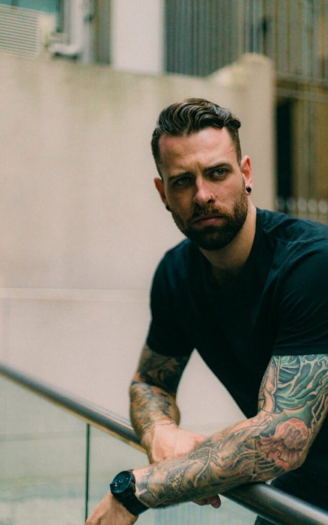 Portrait of a stylish, tattooed man leaning on a railing with a contemplative expression.