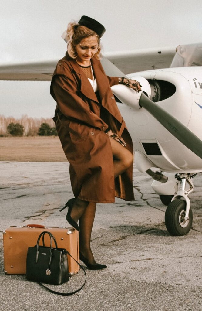 Elegant woman in vintage attire standing by a propeller airplane with luggage on the pavement.