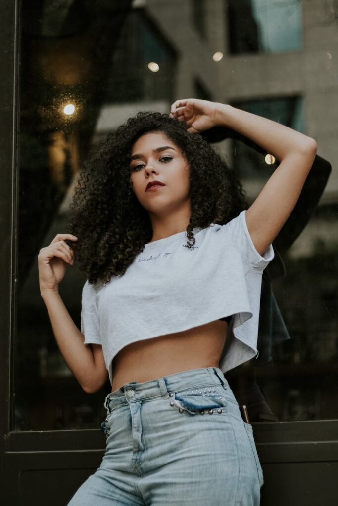 Confident woman with curly hair posing in casual fashion near a glass wall in the city.