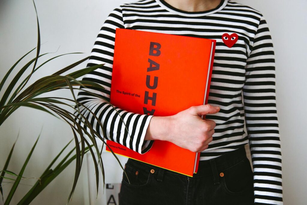 A woman with a striped top holds a Bauhaus book, embracing modern fashion.