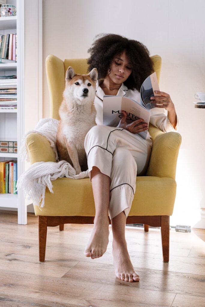 Woman reads magazine with Shiba Inu dog on yellow armchair at home.