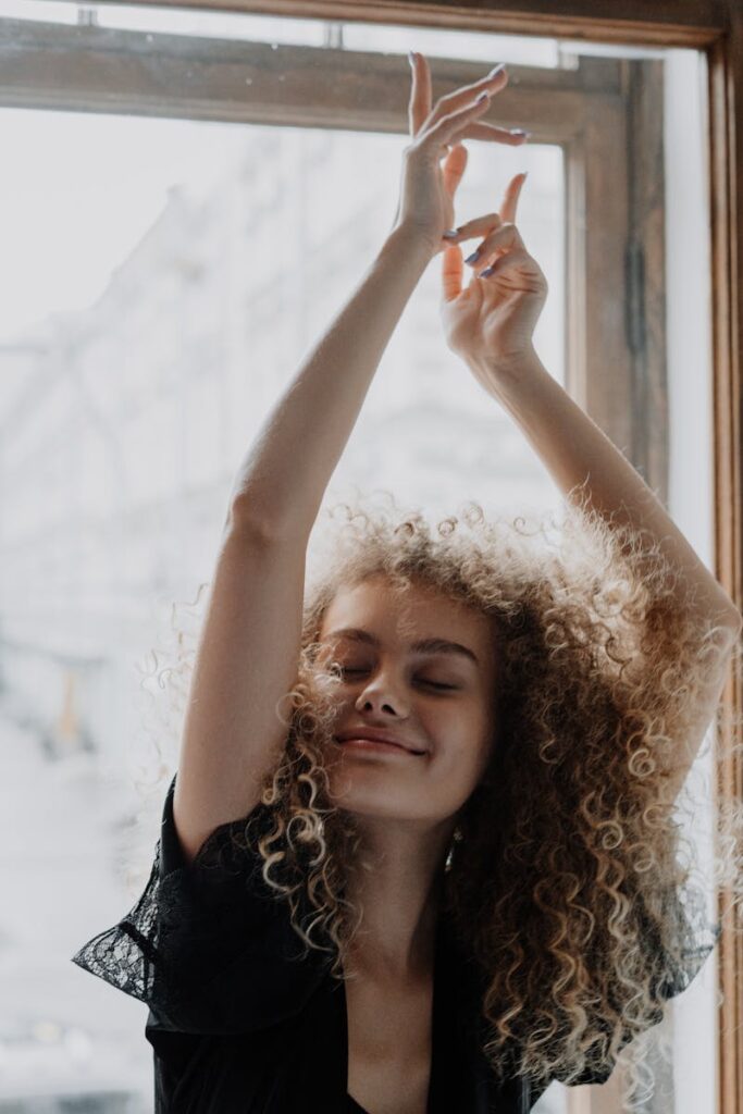 Smiling young woman with curly hair enjoying a moment of joy near a window.