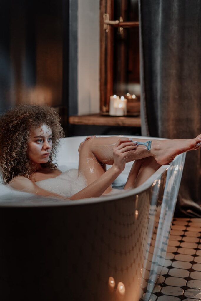 A woman enjoys a relaxing bath with candles while shaving her legs for a tranquil self-care routine.