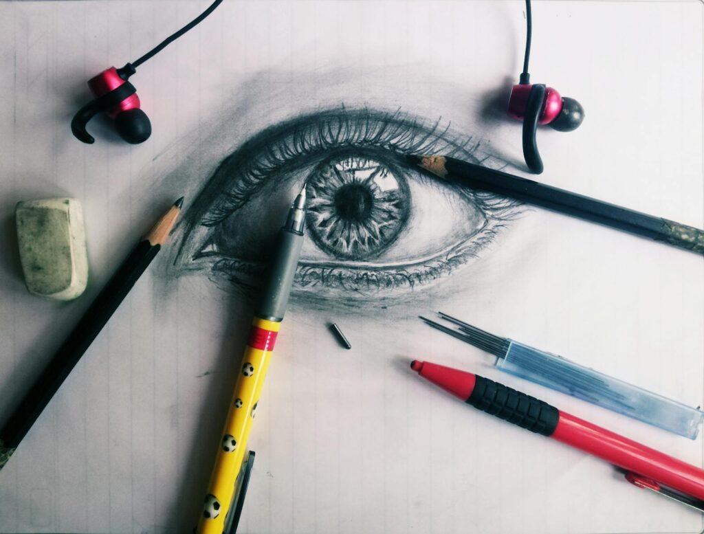 Close-up of a pencil sketch of an eye surrounded by drawing tools and earbuds on paper.
