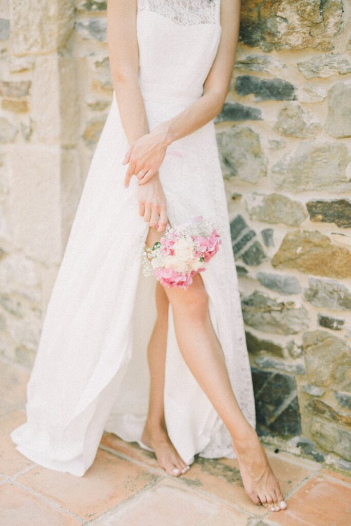 A bride in a serene pose with a bouquet, showcasing a lace wedding dress against a rustic stone wall.