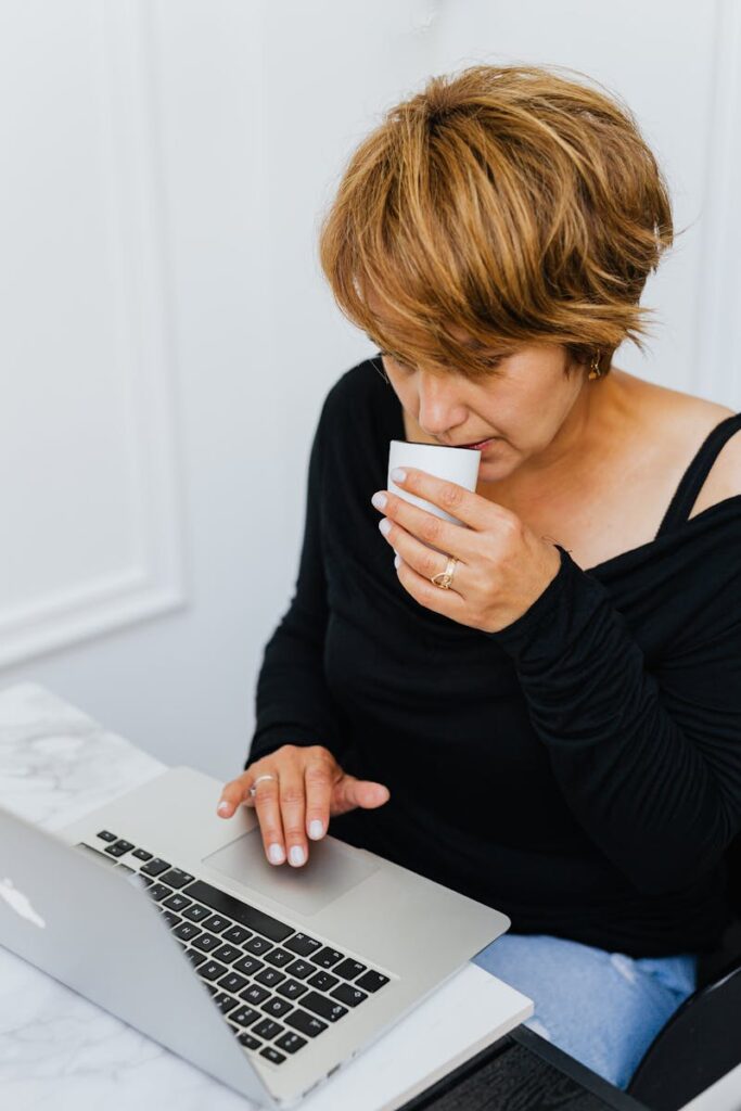 Middle-aged woman in black blouse using laptop and drinking coffee indoors.